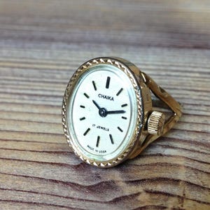 Puede incluir: Un reloj de anillo dorado con una esfera ovalada. La esfera del reloj es blanca con marcas negras y el texto "Chaika 17 Jewels Made in USSR".