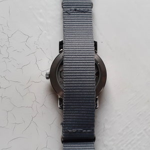 Puede incluir: Un reloj de pulsera con una correa de nailon gris. La esfera del reloj es oscura y la correa es de color gris liso. El reloj est&aacute; sobre un fondo blanco.