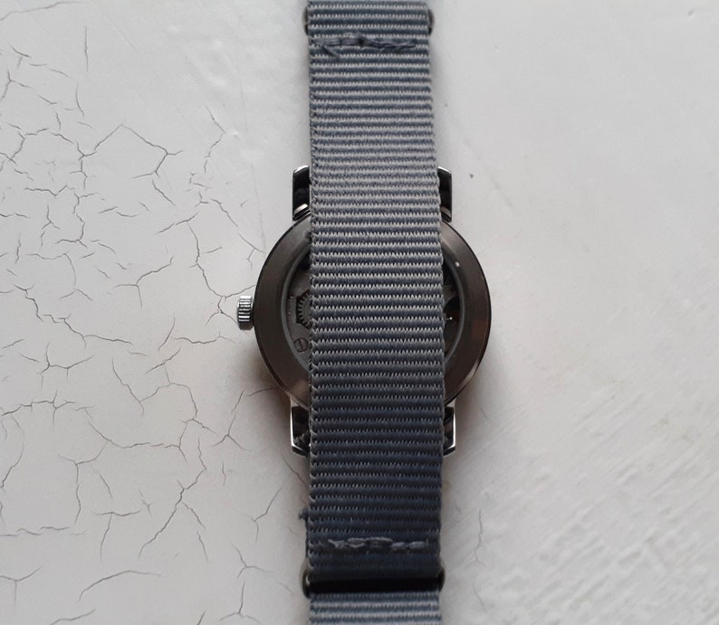 Puede incluir: Un reloj de pulsera con una correa de nailon gris. La esfera del reloj es oscura y la correa es de color gris liso. El reloj est&aacute; sobre un fondo blanco.