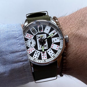 May include: A silver wristwatch with a black face and a green canvas band. The watch face features a design of playing cards with a king of spades in the center.