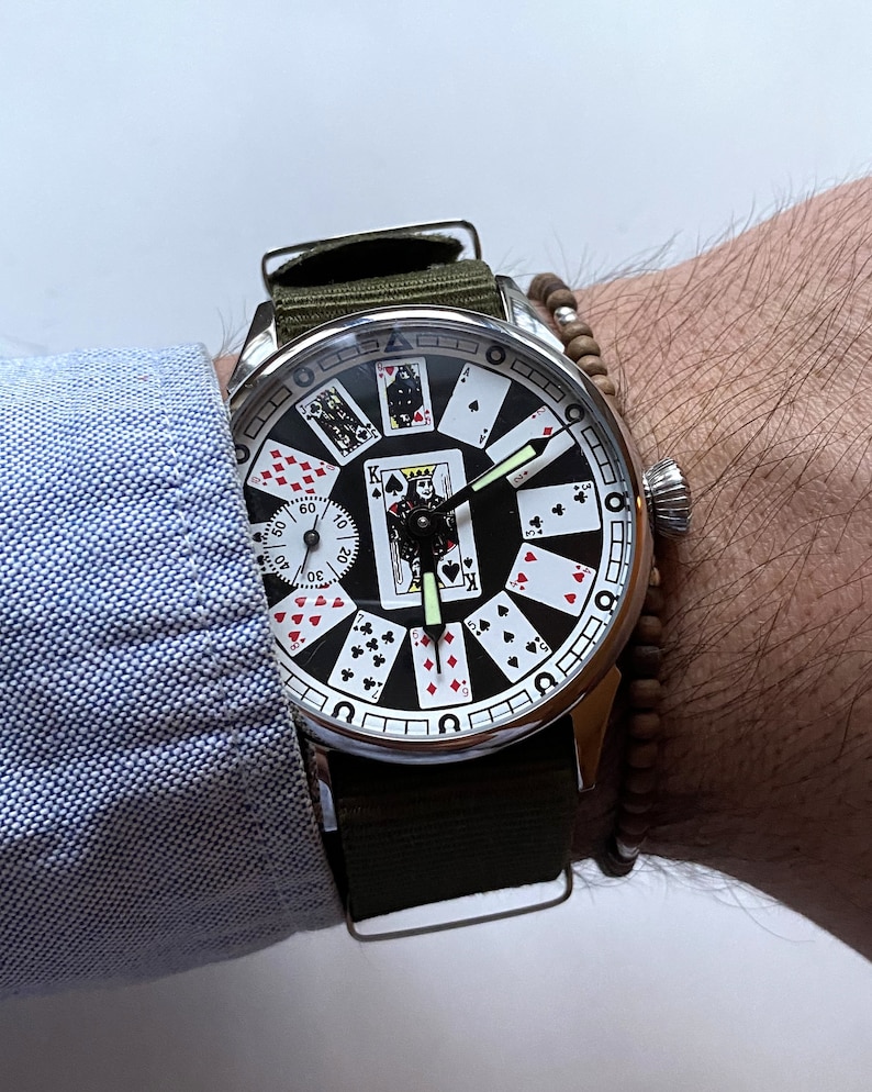 May include: A silver wristwatch with a black face and a green canvas band. The watch face features a design of playing cards with a king of spades in the center.