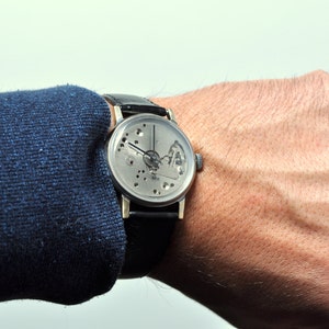 May include: A silver wristwatch with a skeletonized dial and black leather band. The watch is worn on a person's wrist.
