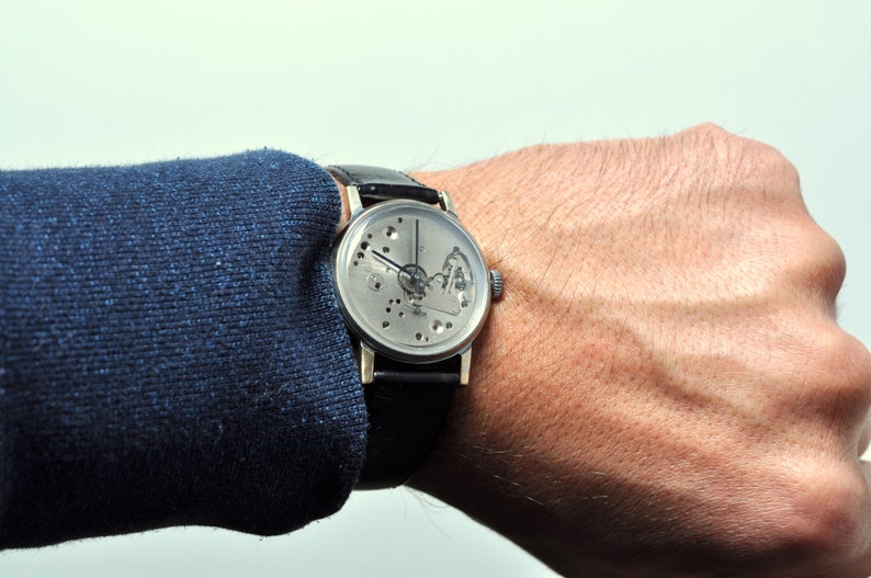May include: A silver wristwatch with a skeletonized dial and black leather band. The watch is worn on a person's wrist.
