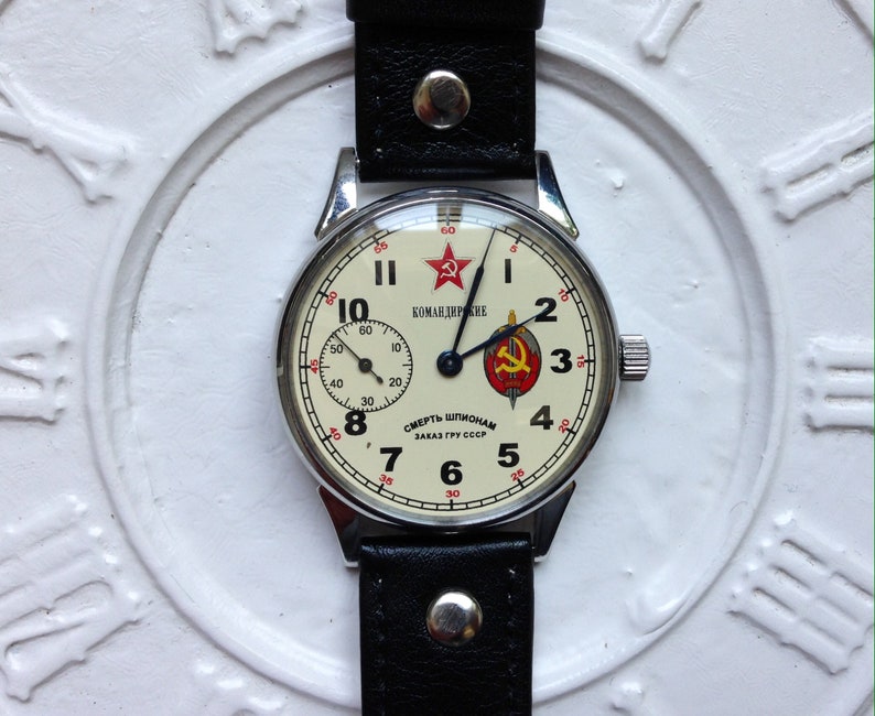 May include: A vintage wristwatch with a white face and black hands. The watch face has red accents and the text "КОМАНДИРСКИЕ" and "СМЕРТЬ ШПИОНАМ ЗАКАЗ ГРУ СССР".