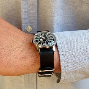 Puede incluir: Un reloj de pulsera plateado con esfera negra y correa de cuero negro. El reloj tiene n&uacute;meros y agujas blancas. Se lleva en la mu&ntilde;eca, con una camisa azul claro visible.