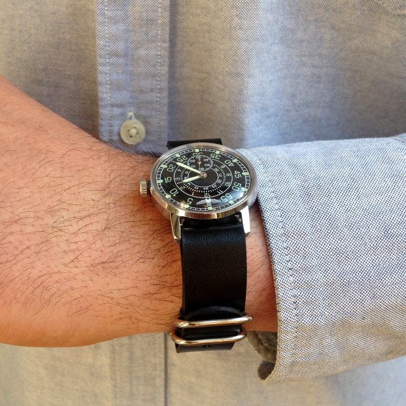 Puede incluir: Un reloj de pulsera plateado con esfera negra y correa de cuero negro. El reloj tiene n&uacute;meros y agujas blancas. Se lleva en la mu&ntilde;eca, con una camisa azul claro visible.
