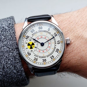 K&ouml;nnte beinhalten: Eine silberne Armbanduhr mit wei&szlig;em Zifferblatt und schwarzem Lederarmband. Das Zifferblatt hat einen schwarzen und blauen Stunden- und Minutenzeiger, einen roten Sekundenzeiger und ein gelbes und schwarzes Strahlungssymbol. Das Zifferblatt der Uhr tr&auml;gt au&szlig;erdem den Text "ВОЙСКА РАДИАЦИОННОЙ, ХИМИЧЕСКОЙ И БАКТЕРИОЛОГИЧЕСКОЙ ЗАЩИТЫ СССР" in kyrillischer Schrift.