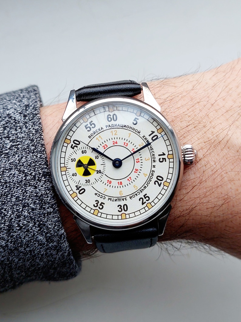 K&ouml;nnte beinhalten: Eine silberne Armbanduhr mit wei&szlig;em Zifferblatt und schwarzem Lederarmband. Das Zifferblatt hat einen schwarzen und blauen Stunden- und Minutenzeiger, einen roten Sekundenzeiger und ein gelbes und schwarzes Strahlungssymbol. Das Zifferblatt der Uhr tr&auml;gt au&szlig;erdem den Text "ВОЙСКА РАДИАЦИОННОЙ, ХИМИЧЕСКОЙ И БАКТЕРИОЛОГИЧЕСКОЙ ЗАЩИТЫ СССР" in kyrillischer Schrift.