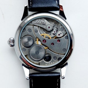 K&ouml;nnte beinhalten: Eine silberne Armbanduhr mit einem schwarzen Lederarmband. Die R&uuml;ckseite der Uhr ist offen und zeigt das komplizierte Uhrwerk.
