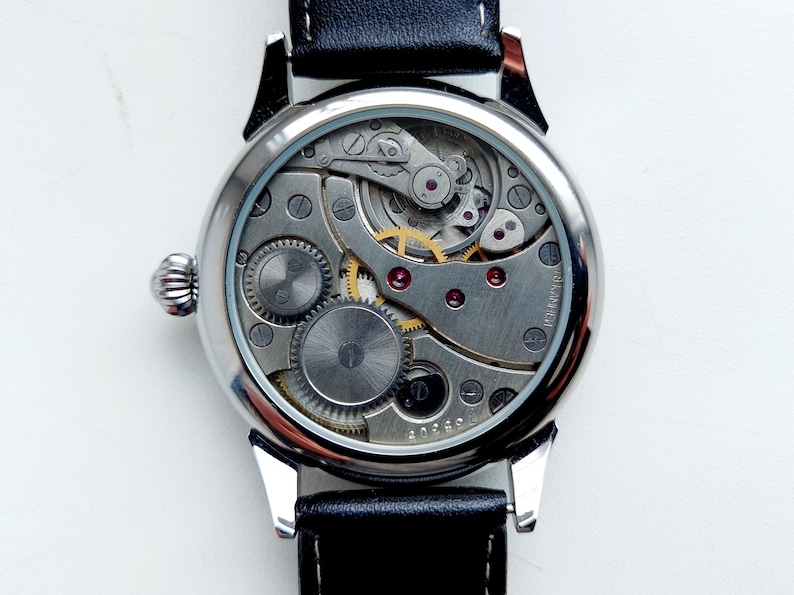 K&ouml;nnte beinhalten: Eine silberne Armbanduhr mit einem schwarzen Lederarmband. Die R&uuml;ckseite der Uhr ist offen und zeigt das komplizierte Uhrwerk.