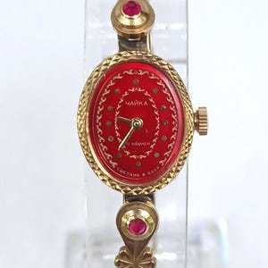 Puede incluir: Un reloj de pulsera vintage dorado con una esfera ovalada roja. El reloj presenta una correa dorada con detalles decorativos y pequeñas piedras preciosas rojas. La esfera del reloj tiene agujas doradas y texto en idioma extranjero.