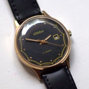 May include: A gold-tone wristwatch with a black face and a black leather band. The watch face has a date window and the text "Raketa 2 Kamen" in Cyrillic.