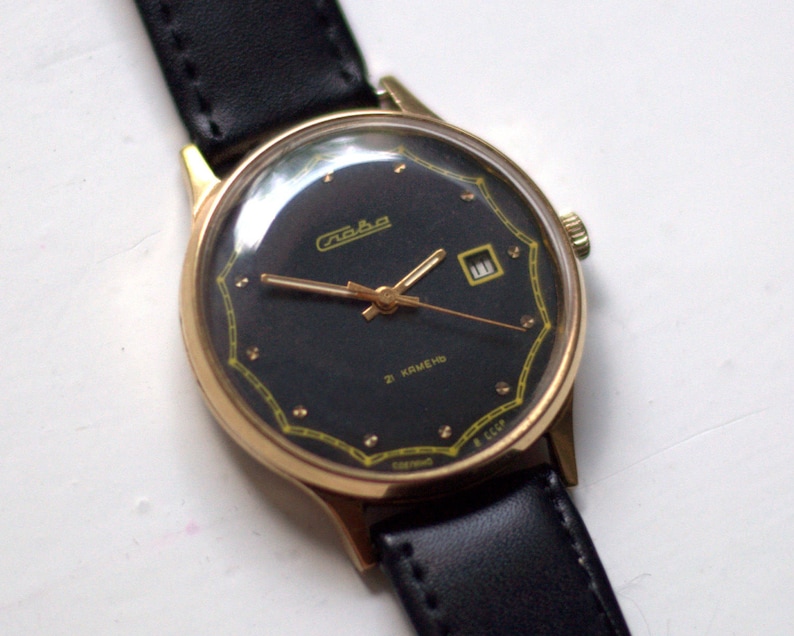 May include: A gold-tone wristwatch with a black face and a black leather band. The watch face has a date window and the text "Raketa 2 Kamen" in Cyrillic.