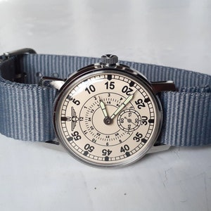 Puede incluir: Un reloj de estilo vintage con esfera crema y n&uacute;meros negros. El reloj tiene una caja plateada y una correa de nailon azul claro. La esfera presenta un peque&ntilde;o dial de segundos y agujas luminosas.