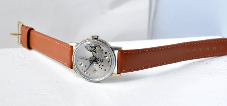 May include: A silver skeleton watch with a brown leather band. The watch face is open, revealing the inner workings of the watch.