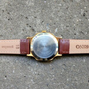 May include: A gold-tone wristwatch with a brown leather band. The back of the watch has the words "TORINO" and "Genuine leather" printed on it. The watch face has the number "220550" printed on it.