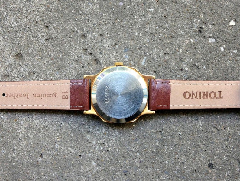 May include: A gold-tone wristwatch with a brown leather band. The back of the watch has the words "TORINO" and "Genuine leather" printed on it. The watch face has the number "220550" printed on it.