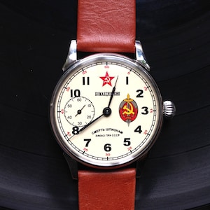 May include: A white wristwatch with a black face and red accents. The watch face features a red star, a hammer and sickle, and the text "КОМАНДИРСКИЕ" and "СМЕРТЬ ШПИОНАМ ЗАКАЗ ГРУ СССР".