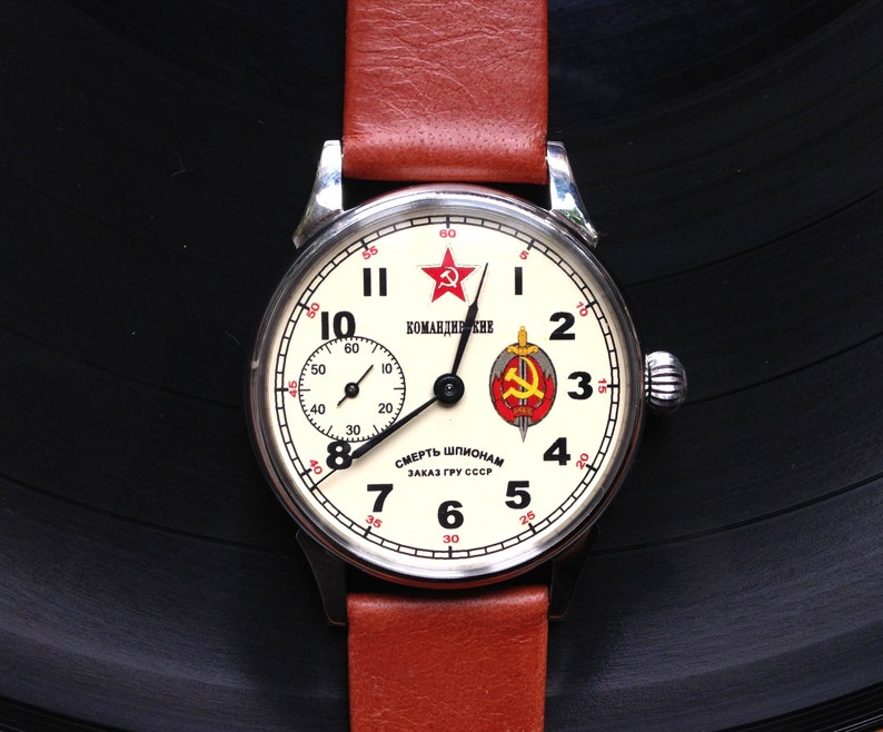 May include: A white wristwatch with a black face and red accents. The watch face features a red star, a hammer and sickle, and the text "КОМАНДИРСКИЕ" and "СМЕРТЬ ШПИОНАМ ЗАКАЗ ГРУ СССР".