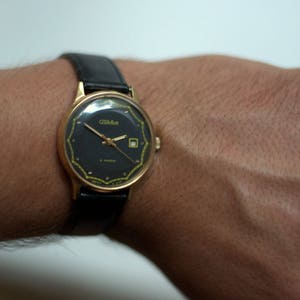 May include: A gold-tone wristwatch with a black face and a black leather band. The watch face has gold-tone hands and markers, and a small date window at the 3 o'clock position. The watch face also has the word "Zlatoust" printed on it.