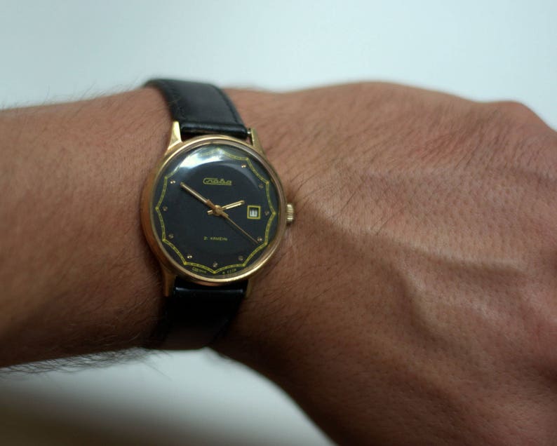 May include: A gold-tone wristwatch with a black face and a black leather band. The watch face has gold-tone hands and markers, and a small date window at the 3 o'clock position. The watch face also has the word "Zlatoust" printed on it.