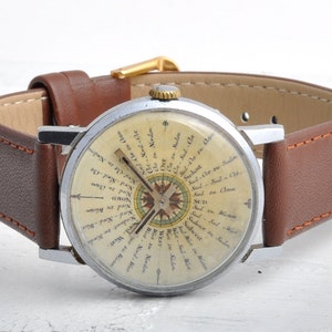 May include: A silver wristwatch with a white face and a brown leather band. The face of the watch features a compass with a detailed compass rose and text in German indicating directions.