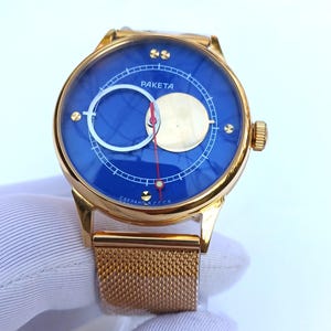 Könnte beinhalten: Eine goldfarbene Armbanduhr mit blauem Zifferblatt und einem Mesh-Armband. Das Zifferblatt hat einen weißen Kreis mit einem kleineren Kreis darin und einen roten Zeiger. Das Zifferblatt der Uhr trägt außerdem den Text "PAKEТА" und "СДЕЛАНО В СССР".