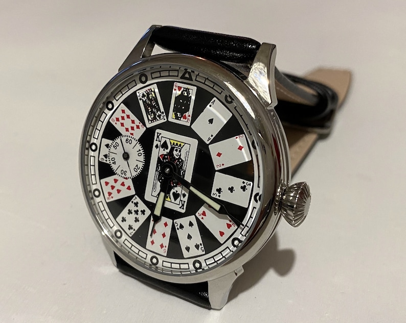 May include: A silver pocket watch with a black face and playing card design. The watch has a black leather band and a silver crown.