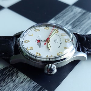 May include: A vintage wristwatch with a silver case, a cream-colored face, and a black leather band. The watch face features gold numerals, red and gold hands, and a logo. The watch is displayed on a black and white checkered surface.