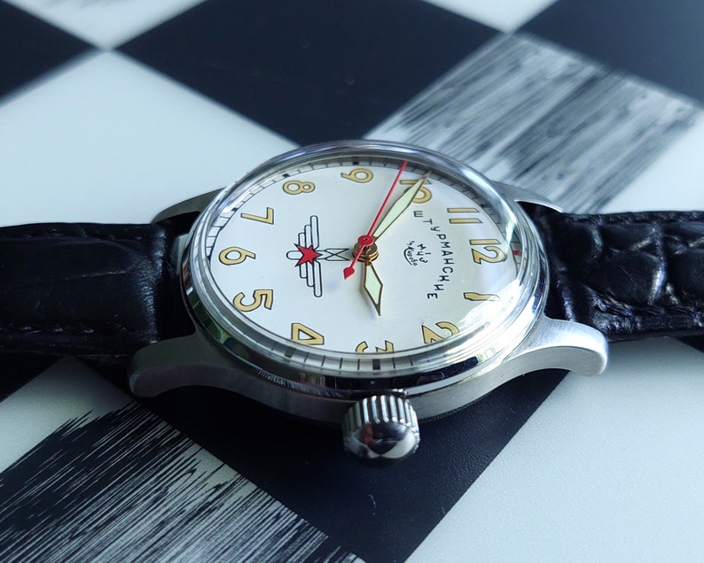 May include: A vintage wristwatch with a silver case, a cream-colored face, and a black leather band. The watch face features gold numerals, red and gold hands, and a logo. The watch is displayed on a black and white checkered surface.