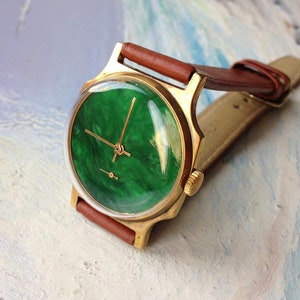 May include: A gold-tone wristwatch with a green, marbled face and brown leather band.