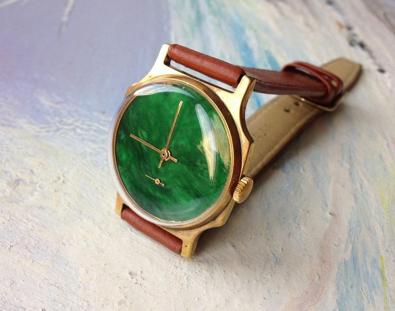 May include: A gold-tone wristwatch with a green, marbled face and brown leather band.