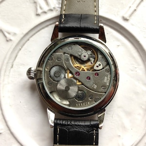 May include: A silver watch with a black leather band. The back of the watch is clear, revealing the intricate mechanical workings of the watch.