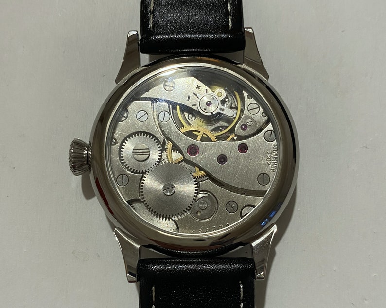 May include: A silver wristwatch with a black leather band. The watch face is clear and shows the inner workings of the watch.
