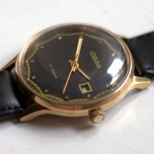 May include: A gold-tone wristwatch with a black face and a black leather band. The watch face has a gold-tone border and a small date window at the 3 o'clock position. The watch face also has the word "Chaika" printed in gold letters.