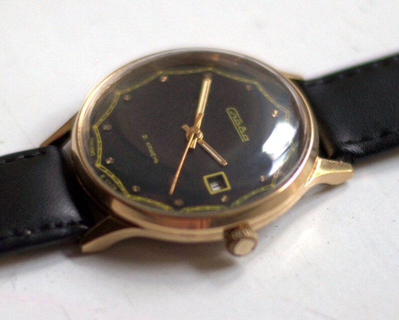 May include: A gold-tone wristwatch with a black face and a black leather band. The watch face has a gold-tone border and a small date window at the 3 o'clock position. The watch face also has the word "Chaika" printed in gold letters.