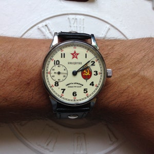 May include: A silver wristwatch with a white face and black leather band. The face has black Roman numeral hour markers, black hands, and a red star with a hammer and sickle. The text on the face reads "Командирские" and "Смерть Шпионам ЗАКАЗ ГРУ СССР".