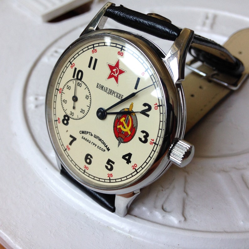 May include: A silver wristwatch with a white face and black hands. The watch face has red accents and the text "КОМАНДИРСКИЕ" and "СМЕРТЬ ШПИОНАМ ЗАКАЗ ГРУ СССР".