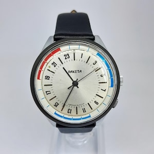 Peut inclure: Une montre-bracelet Raketa avec un cadran argenté affichant des marquages de 24 heures, des aiguilles noires et un bracelet en cuir noir. Le cadran présente des sections rouge et bleu sur le bord et le texte PAKETA et СДЕЛАНО В СССР.