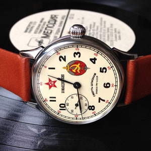May include: A silver wristwatch with a white face and black hands. The watch face has a red star and the words "KOMANDIRSKIE" and "СМЕРТЬ ШПИОНАМ ЗАКАЗ ГРУ СССР". The watch has a brown leather band.