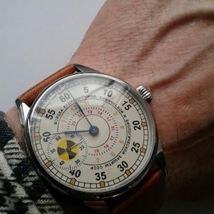 K&ouml;nnte beinhalten: Eine Armbanduhr mit wei&szlig;em Zifferblatt und schwarzen Zeigern. Die Uhr hat ein braunes Lederarmband und ein silbernes Geh&auml;use. Das Zifferblatt der Uhr hat ein rotes und schwarzes Strahlungssymbol und den Text "ВОЙСКА РАДИЦИОННОЙ, ХИМИЧЕСКОЙ И БИОЛОГИЧЕСКОЙ ЗАЩИТЫ СССР".