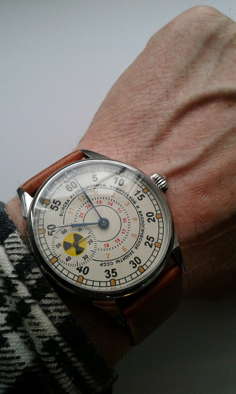 K&ouml;nnte beinhalten: Eine Armbanduhr mit wei&szlig;em Zifferblatt und schwarzen Zeigern. Die Uhr hat ein braunes Lederarmband und ein silbernes Geh&auml;use. Das Zifferblatt der Uhr hat ein rotes und schwarzes Strahlungssymbol und den Text "ВОЙСКА РАДИЦИОННОЙ, ХИМИЧЕСКОЙ И БИОЛОГИЧЕСКОЙ ЗАЩИТЫ СССР".