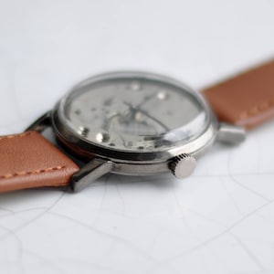 May include: A silver wristwatch with a brown leather band. The watch face is clear and shows the inner workings of the watch.