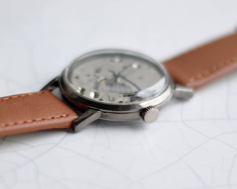 May include: A silver wristwatch with a brown leather band. The watch face is clear and shows the inner workings of the watch.