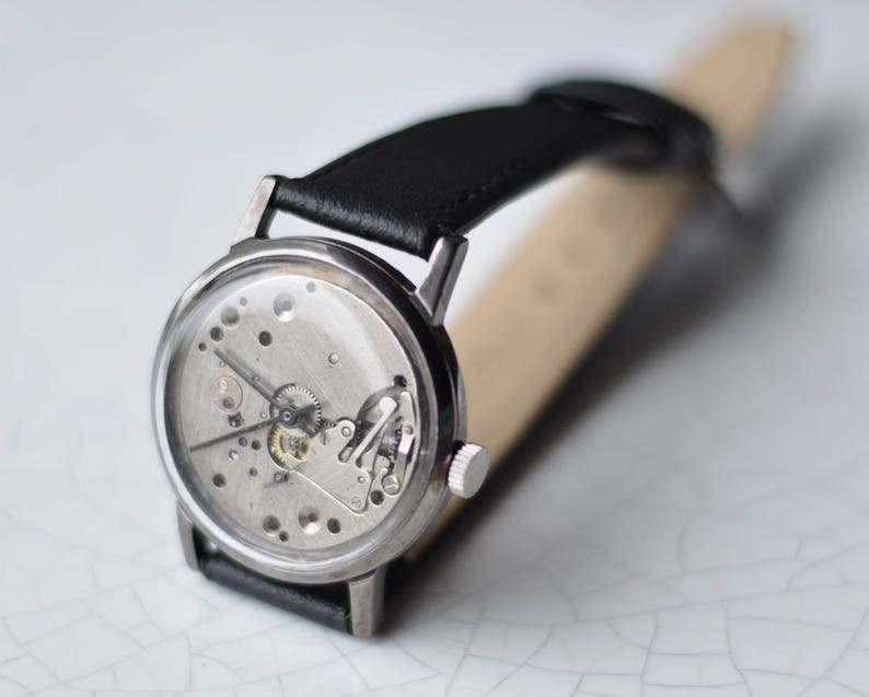 May include: A silver wristwatch with a black leather band. The watch face is open, revealing the inner workings of the watch.
