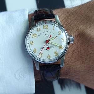 May include: A silver-toned wristwatch with a cream-colored face and black leather band. The watch face features gold-colored numerals, red and black hands, and the text "Штурманские". The watch is on a wrist with a white shirt cuff.