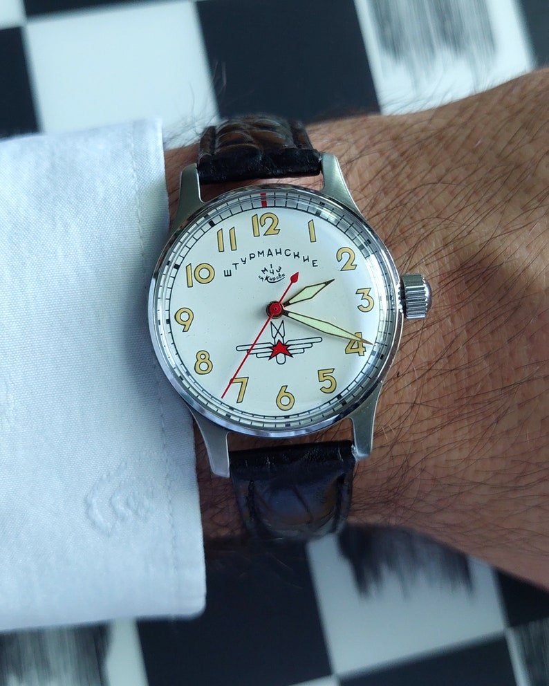 May include: A silver-toned wristwatch with a cream-colored face and black leather band. The watch face features gold-colored numerals, red and black hands, and the text "Штурманские". The watch is on a wrist with a white shirt cuff.
