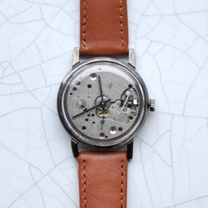 May include: A silver wristwatch with a brown leather band. The watch face is exposed, showing the inner workings of the watch.