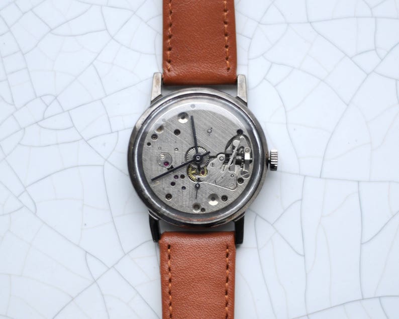 May include: A silver wristwatch with a brown leather band. The watch face is exposed, showing the inner workings of the watch.