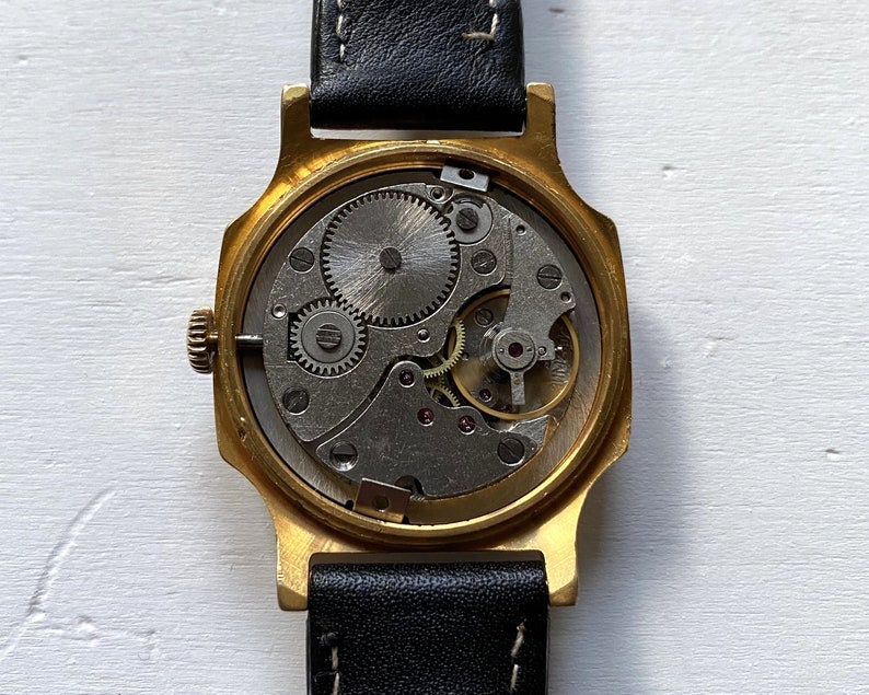 May include: A gold-toned wristwatch with a black leather band. The back of the watch is open, revealing the intricate mechanical workings of the watch.
